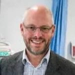 A photo of Dr Alan Whone. A man with short grey hair, a beard and glasses in a hospital setting. 
