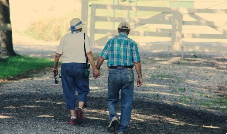 An older woman and older man wearing jeans shirts and hats walking along a gravel path on a sunny day, holding hands. 