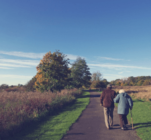 Photo of an older man and woman linked arms, walking down a path. Trees and blue sky in the background. 