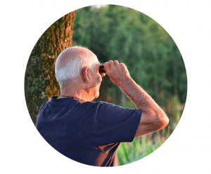 Circle cropped photo of an older man with grey hair looking through binoculars. A tree trunk and trees are in the background. 