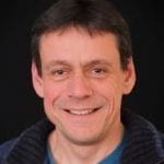 A photo of Prof Will Hollingworth. A man with short dark hair against a black background. 